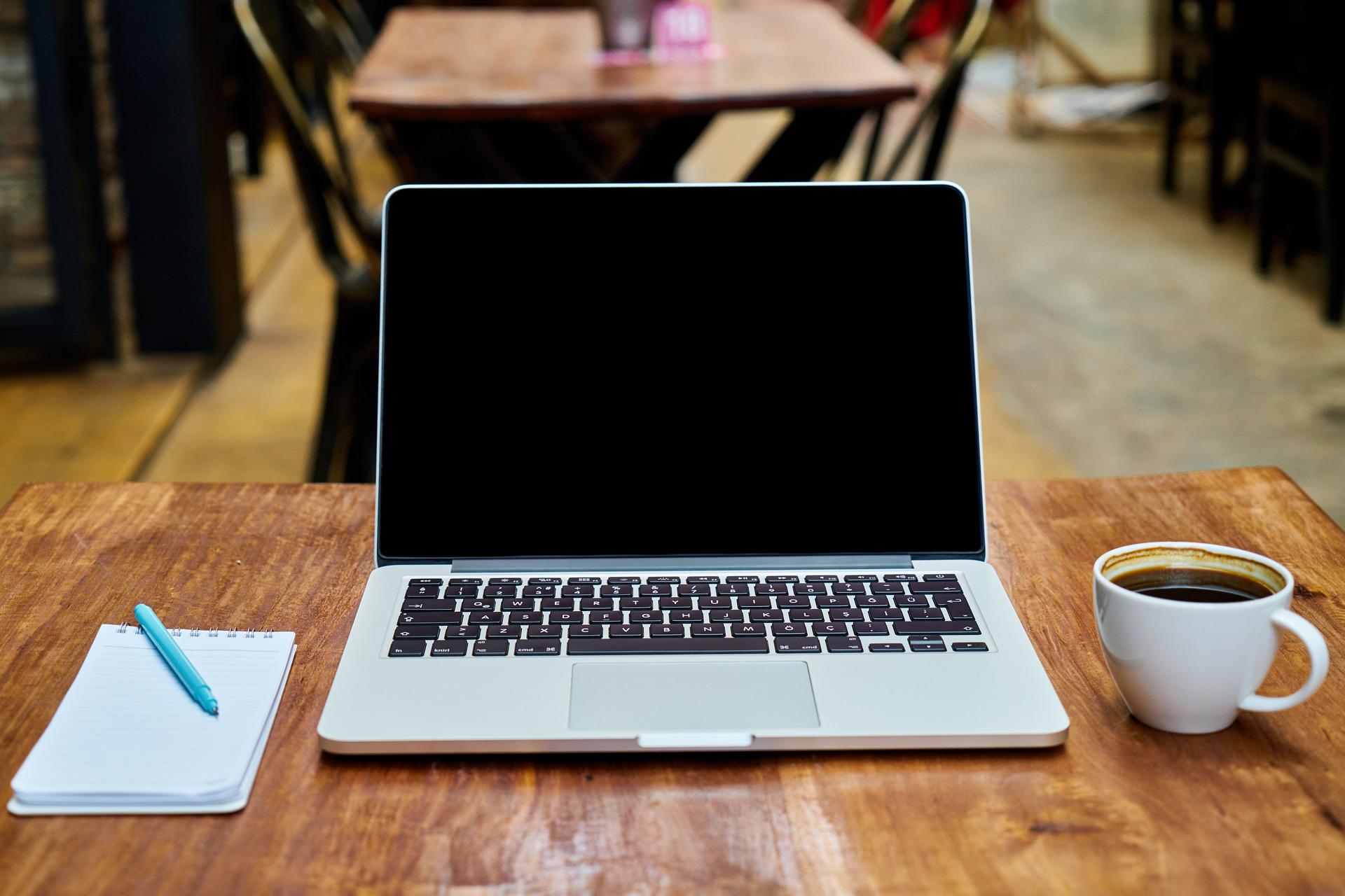 An open laptop, with a notebook and pen on the left and a coffee mug on the right.