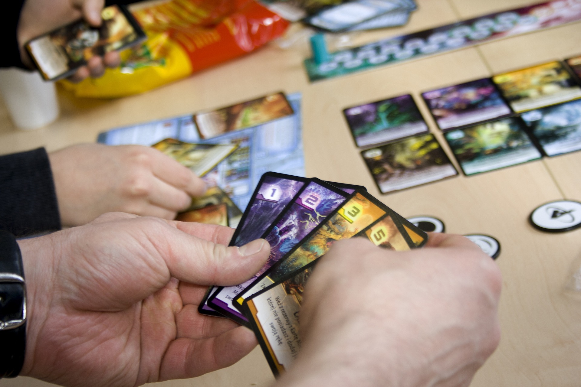 A set of hands holding cards in front of a gaming set up with snacks in the background.