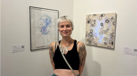 Photo of the artist Jessica Pixie in front of 2 of her pieces, a painting and a textile installation
