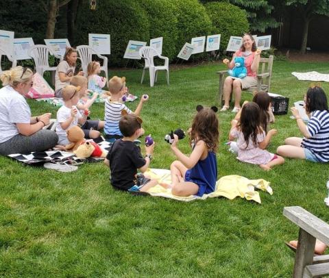 Kids and their families sit on picnic blankets outside and listen to a teddy bear story.