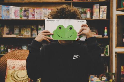 A child in a library holding up a book with a frog on it that they are reading.