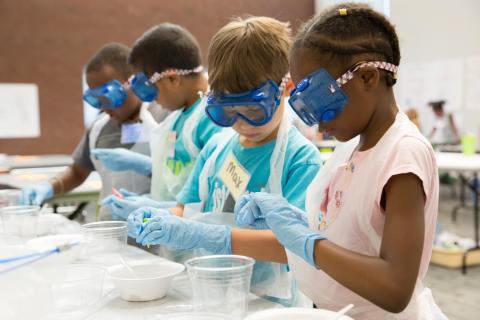 Multicultural group of children wearing goggles and rubber gloves complete a science experiment.