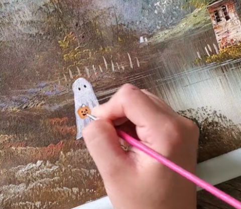 A hand of a person painting a white-sheet style ghost into a landscape painting.