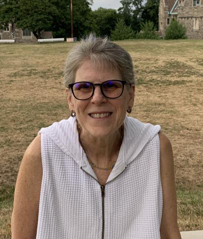 Photo of artist Carol Beth Yoffee, an older white woman with short grey hair wearing glasses and a sleeveless white top