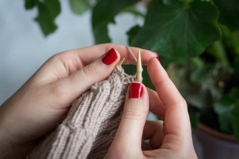 A close up photo of hands in red nail polish knitting with tan yarn.