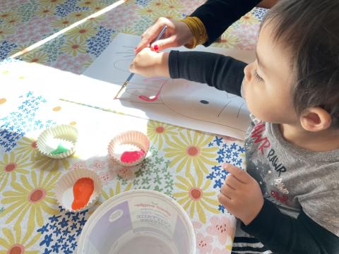 A mother's hand guides her young son as he paints with a paintbrush on paper.