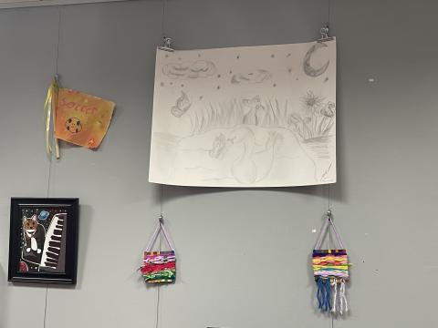 Photo of some of the art in the youth art show: a textile flag, a drawing of a cat playing the piano, a pencil sketch of a swan, and rainbow weavings