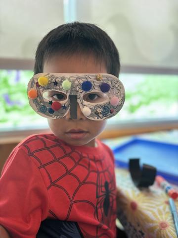 A child in a Spiderman costume wears a paper mask covered in pom poms.
