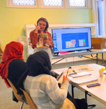 Photo of several hijab wearing women around a table, an ESL instructor stands in the front of the room with lessons on a screen