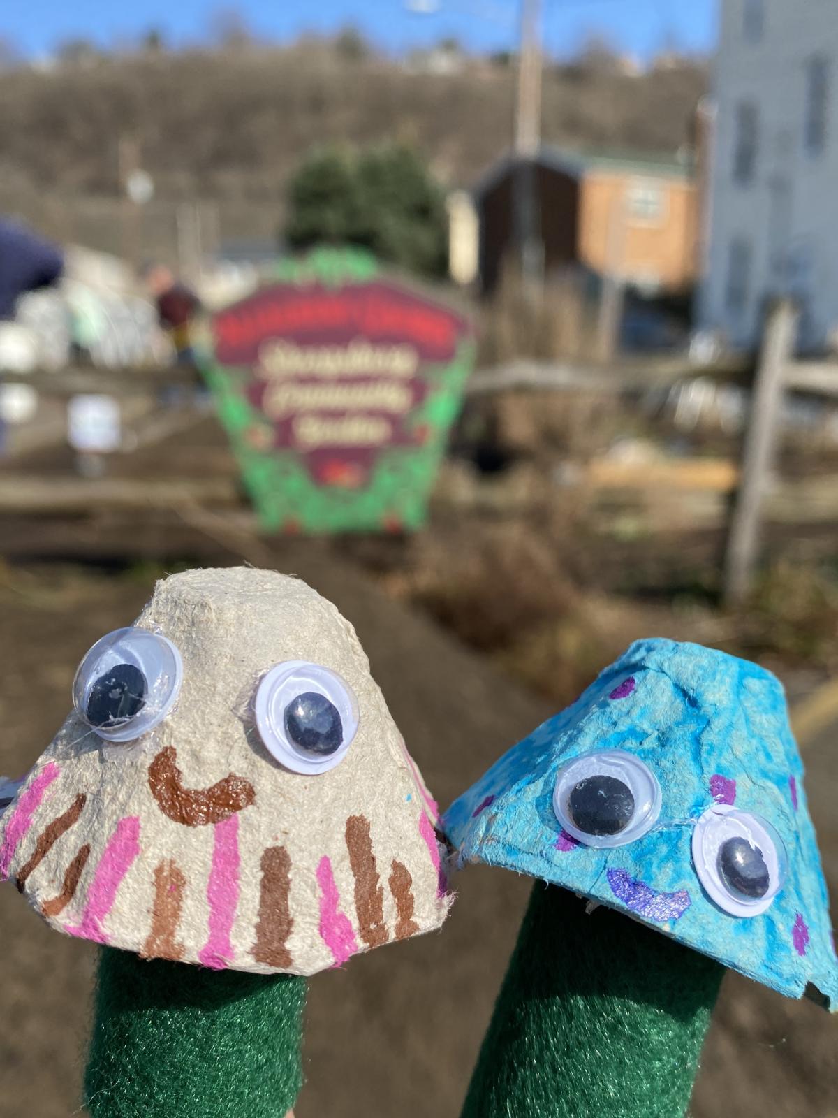 Two mushroom finger puppets with google eyes pose in front of the garden.
