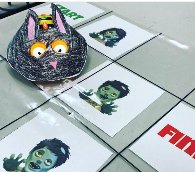 A Bee Bot is wearing a paper, black cat costume and navigating a game board with vampires.