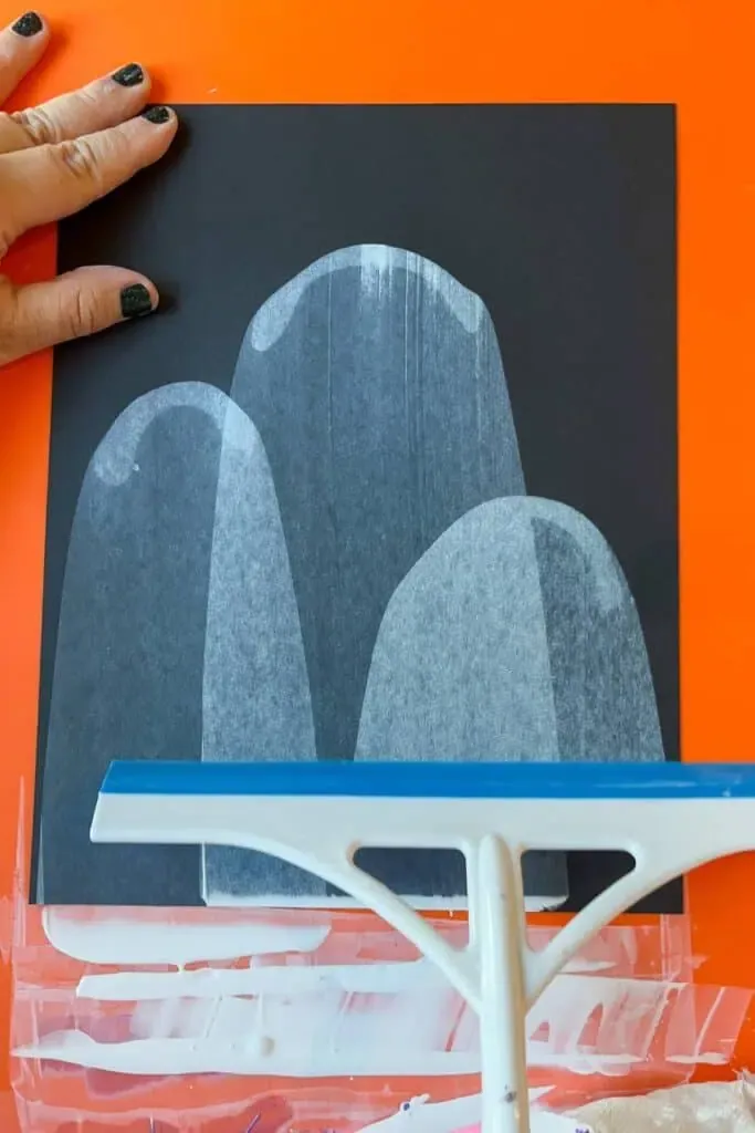 A child's hand drags white paint across black paper with a squeegee.