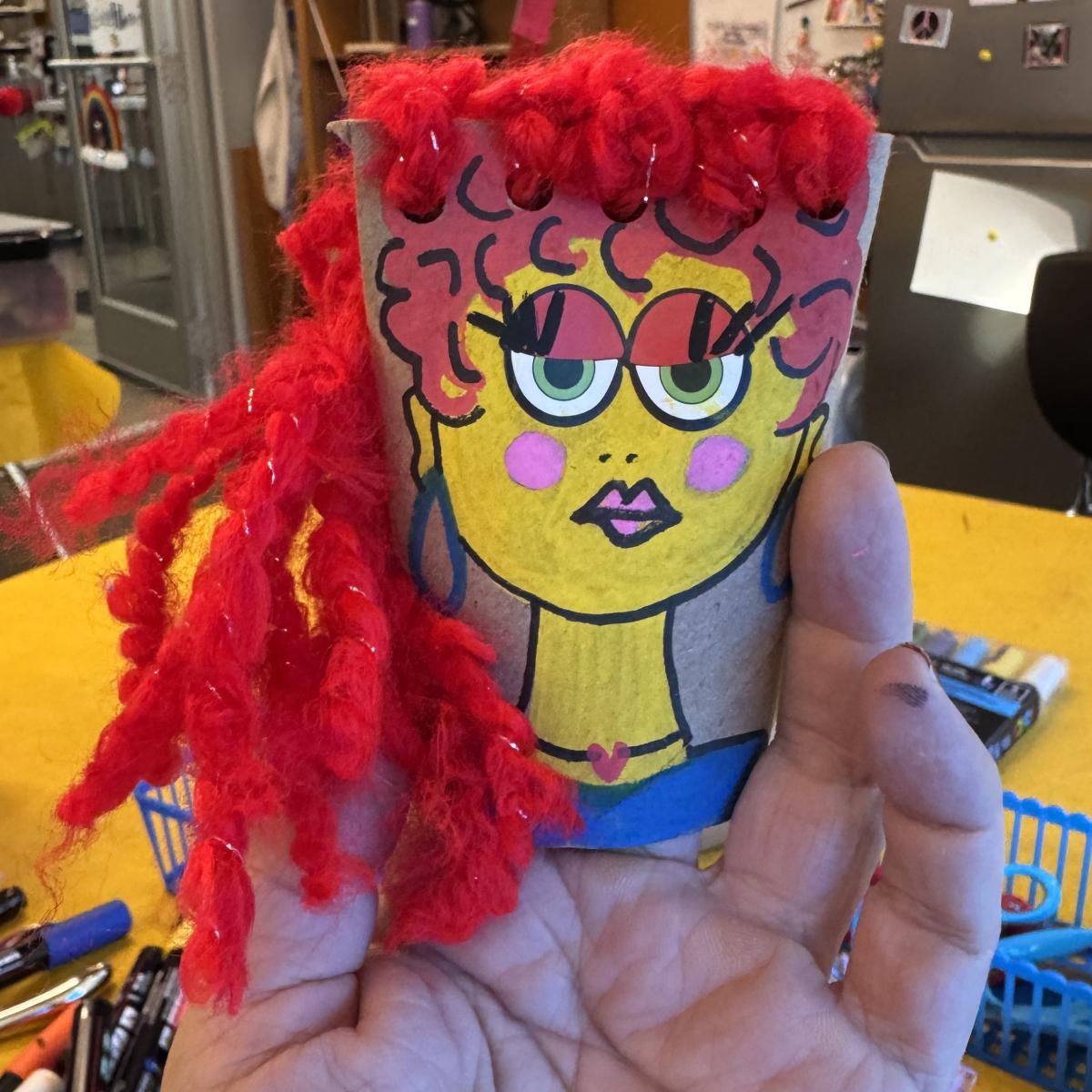 A puppet created from a toilet paper roll features a smiling woman with red hair.